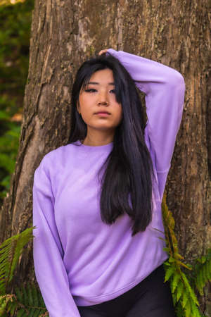 Portrait of a young Asian brunette in a park next to a tree with a pink sweater.の写真素材