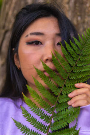 Portrait of a young Asian brunette in a park next to a tree and a fern leaf near her face.の写真素材