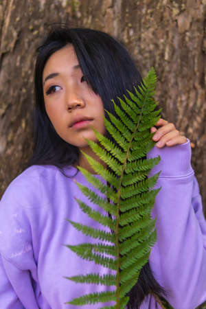 Portrait of a young Asian brunette in a park next to a tree and a fern leaf near her face.の写真素材