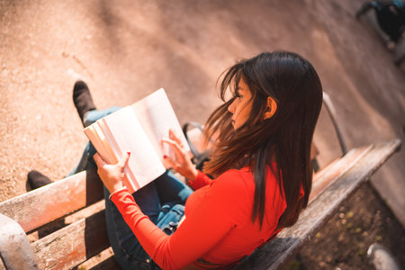 Lifestyle. Young Caucasian brunette in a red shirt sitting on a bench in the park reading a book....