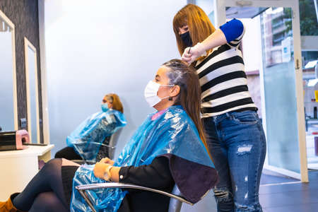 Opening of hairdressers in the coronavirus pandemic, covid-19. Security measures, mask, protective screen, social distance. Applying dye to clients in the new normalの写真素材