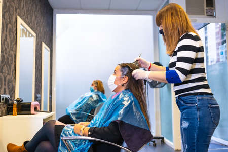 Opening of hairdressers in the coronavirus pandemic, covid-19. Security measures, mask, protective screen, social distance. Applying dye to clients in the new normalの写真素材