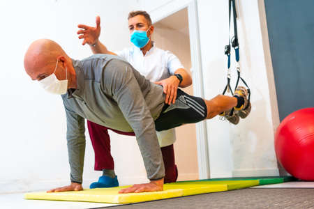Physiotherapist with a mask and a patient doing push-ups with their feet on rubber bands. Physiotherapy with protective measures for the Coronavirus pandemic, COVID-19. Osteopathy, sports quiromassageの写真素材