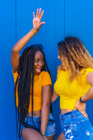 Lifestyle, very happy girl friends. Black girl with long braids and blonde Caucasian girl. Dressed in yellow on a blue backgroundの写真素材