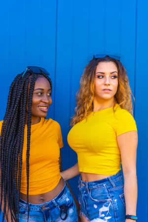 Lifestyle, very happy teenage girl friends on a blue wall background dressed in yellow t-shirts. Black girl with long braids and blonde Caucasian girl.の写真素材