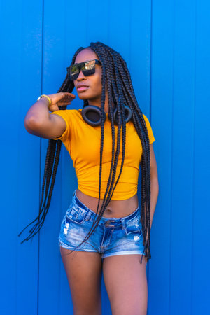 Lifestyle, black girl with long braids, in a yellow shirt and sunglasses on a blue background. Dj...