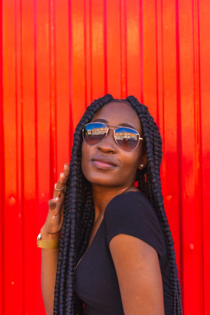 Lifestyle, black girl with big braid in black t-shirt, denim shorts and sunglasses on a red background. Portrait of a young trap dancerの写真素材