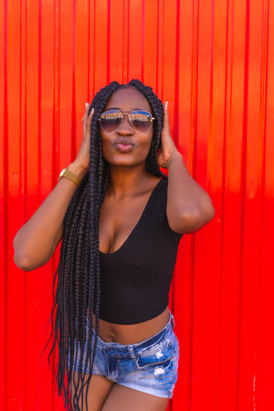 Lifestyle, black girl with big braid in black t-shirt, denim shorts and sunglasses on a red...