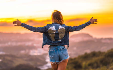 Lifestyle, a Caucasian girl with a denim jacket and an angel wings print on the back, with denim shorts. With open arms atop a mountain in a gorgeous summer sunsetの写真素材