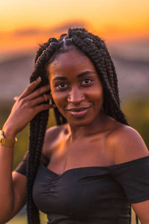 Lifestyle, Latina girl with black skin with beautiful long braids in her hair, wearing a tight black short dress. Portrait of the young man on top of the mountain in a beautiful summer sunsetの写真素材