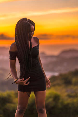 Lifestyle, Latina girl with black skin with beautiful long braids in her hair, wearing a tight black short dress. Portrait of the young man on top of the mountain in a beautiful summer sunsetの写真素材
