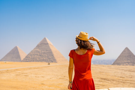A young tourist in a red dress looking at the Pyramids of Giza, the oldest Funerary monument in the world. In the city of Cairo, Egyptの写真素材