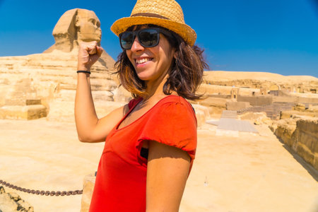 A young tourist in a red dress joking at the Great Sphinx of Giza and in the background the Pyramids of Giza, the oldest Funerary monument in the world. In the city of Cairo, Egyptの写真素材