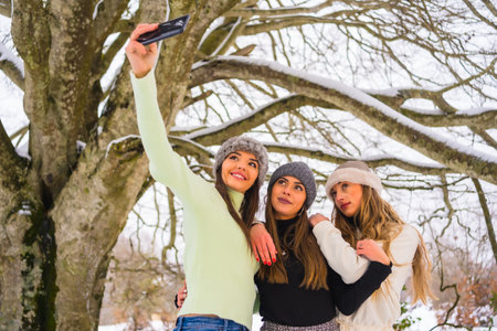 Winter lifestyle, three beautiful caucasian friends taking a selfie with the phone in the snow under a tree, holidays in natureの写真素材