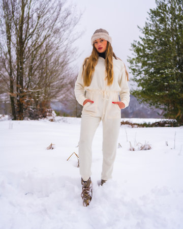 Winter lifestyle, portrait of young pretty blond Caucasian woman with white winter outfit and woolen hat in snow next to snowy pine trees, vacation in natureの写真素材