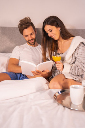 A couple in pajamas reading a book in the breakfast of coffee and orange juice in the hotel bed, lifestyle of a couple in love. Vertical photoの写真素材