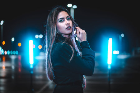 Caucasian brunette in pink wool jacket in an empty car park illuminated with blue light. Night urban session in the cityの写真素材