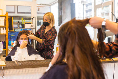 Blonde hairdresser with face protection mask performing a hairstyle to the client. Security measures in the Covid-19 pandemic, vertical photoの写真素材