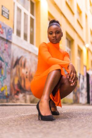 Fashionable urban style with a black African girl in an orange dress and black heels on a city street. Crouched poseの写真素材