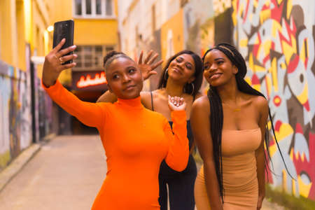 Three girls taking a selfie. Fashionable urban style with three black African girls on a city street, tight dresses. Girlfriends lifestyleの写真素材