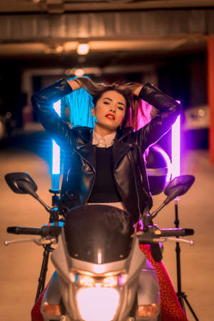 Photography with blue and pink neons on a motorcycle. Portrait of a young pretty blond Caucasian woman in a black leather jacketの写真素材