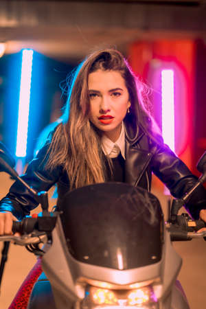 Photography with blue and pink neons on a motorcycle. Portrait of a young pretty blond Caucasian woman in a black leather jacketの写真素材