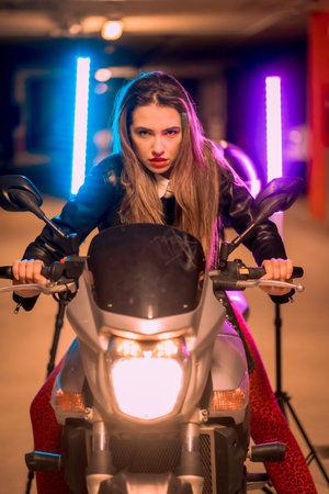 Photography with blue and pink neons on a motorcycle. Portrait of a young pretty blond Caucasian woman in a black leather jacketの写真素材