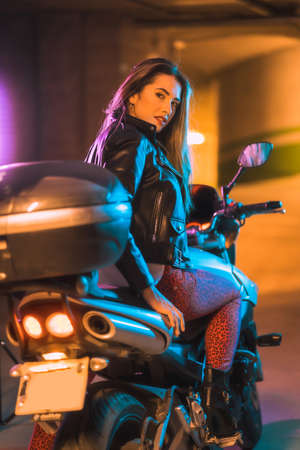 Photography with blue and pink neons on a motorcycle from behind. Portrait of a young blonde Caucasian model wearing a black leather jacketの写真素材