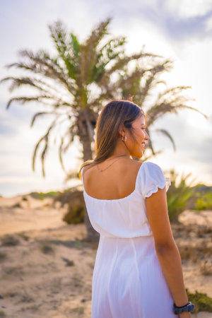 Lifestyle of a young Caucasian brunette enjoying the beach vacation next to a palm tree in a white dress, vertical photoの写真素材