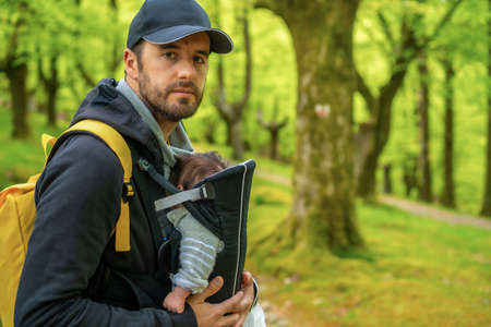 A young father with a yellow backpack and a black cap walking with the newborn child in the backpack on a path in the woodsの写真素材