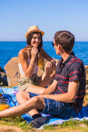A young Caucasian couple eating some ham at the picnic in the mountains by the sea enjoying the heat, summer lifestyleの写真素材