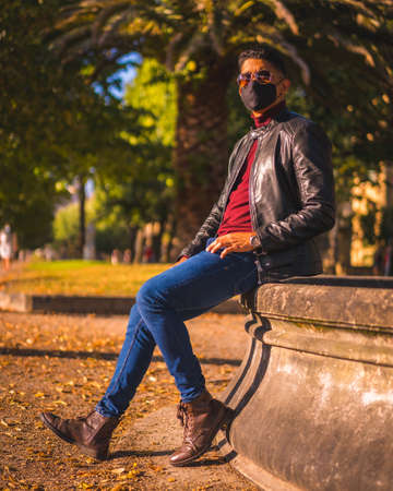Fashion lifestyle, portrait of a young Latino in the city at a water fountain. Jeans, leather jacket and brown shoes. In a pandemic with a maskの写真素材