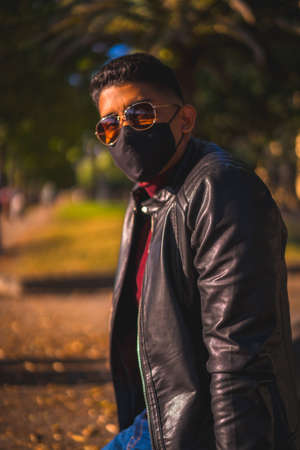 Fashion lifestyle, portrait of a young Latino in the city at a water fountain. Jeans, leather jacket and brown shoes. In a pandemic with a maskの写真素材