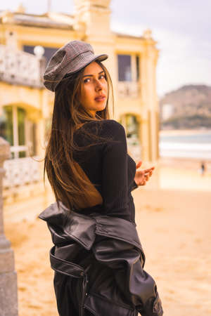 Lifestyle, portrait of a young brunette Caucasian woman sitting on some stairs in a short black dress, with a beret and plaid stockings next to a beach, vertical photoの写真素材