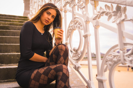 Lifestyle in the city, Caucasian brunette in a short black dress and with plaid tights sitting...