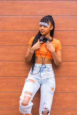Lifestyle with a young trap dancer in the city. Black grind girl of African ethnic group with orange t-shirt and cowboy pants, with headphones on a wooden backgroundの写真素材