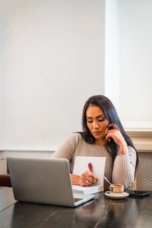Brunette girl teleworking in a cafeteria with laptop, taking notes in online meeting, vertical photoの写真素材