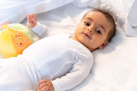5 month old Caucasian baby smiling in white bed just woken up, white bodysuit and a ball to playの写真素材
