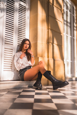 Lifestyle, a young brunette in a black leather skirt and a white shirt sitting next to a white blind of a traditional house in autumnの写真素材