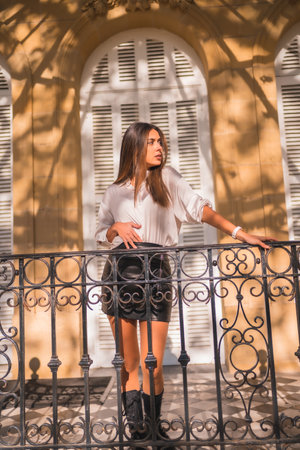 Lifestyle, sweet look of a young brunette in a black leather skirt and a white shirt on a black railing of a traditional houseの写真素材