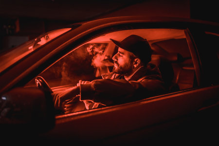 Man in a cap driving car smoking at night in a garage lit with a red light, sports carの写真素材