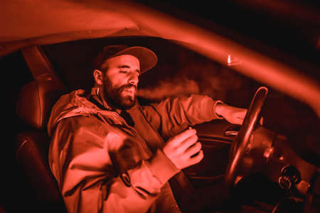 Rapper man in a cap in a car smoking at night in a garage lit with a red light in winter, sports carの写真素材