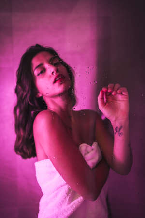 Lifestyle, sexy brunette Caucasian girl with towel at the end of the shower with sensual look, with pink led light, portraits inside the wet showerの写真素材