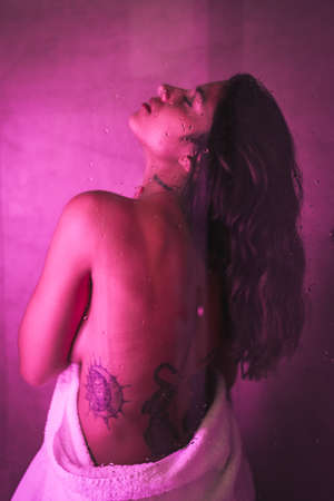 Lifestyle, sexy brunette Caucasian girl with towel at the end of the shower with sensual gaze, with pink led light, portraits inside the shower with bare backの写真素材