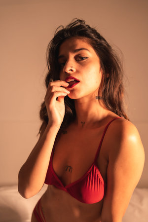 Lifestyle, sexy brunette Caucasian girl with a red tight corset, sensual perched on the bed, illuminated with dim light at night in the roomの写真素材