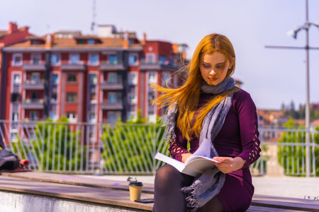 Beautiful woman in purple dress and scarf reading a book in a park in the city, imagining and...