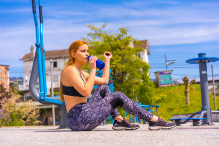 Red-haired woman with purple leggings and black overalls exercising on the machines, drinking water on a break, sport in the cityの写真素材