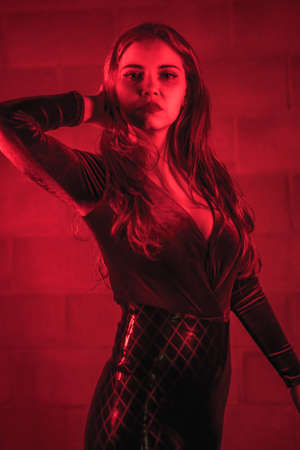 Red-haired girl in latex, brown and black tight suit on a brick wall, posed at night illuminated with red lightの写真素材