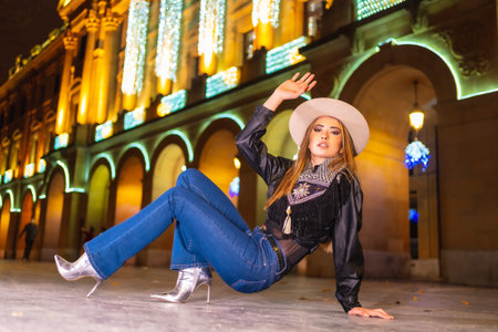 Young caucasian blonde in jeans, hat and a black jacket in the illuminated city at christmas, perched on a ground crouchingの写真素材