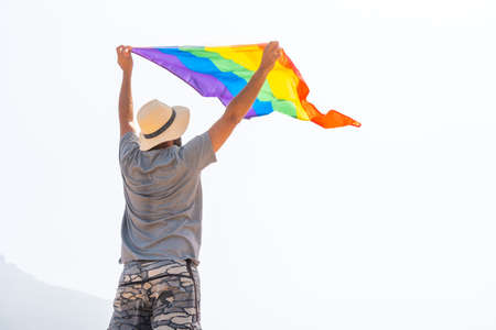 An unrecognizable gay person in a gray t-shirt and white hat waving the LGBT flag in a cloudy sky, symbol of homosexualityの写真素材